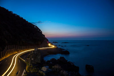 Light trail on road by sea against sky at dusk