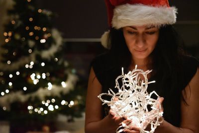 Woman holding illuminated lights against christmas tree at home