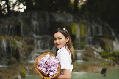 Portrait of a smiling young woman holding bouquet of flowers while standing against waterfall