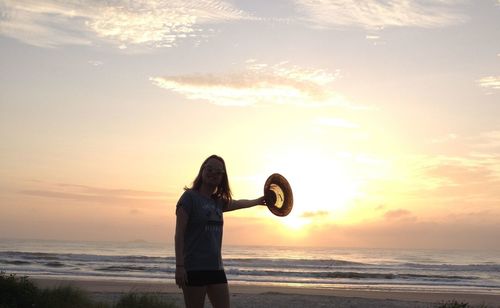 Portrait of young woman holding hat while standing at beach against sky during sunset