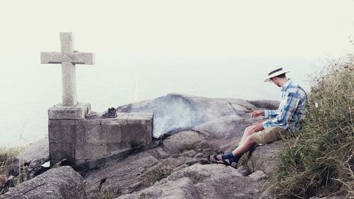 Man sitting by concrete cross on mountain against clear sky