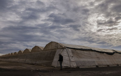 Adult man standing in front of plastic greenhouses against cloudy sky in almeria, spain