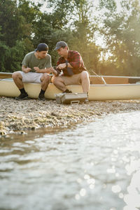 Male friends adjusting fishing rods while sitting on boat by lake