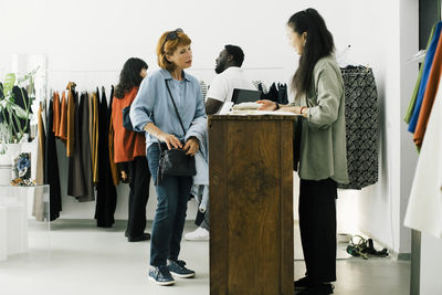 Senior woman discussing with female sales staff while standing at checkout counter in clothes store