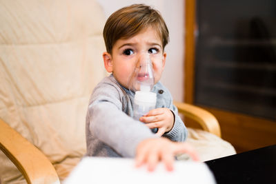 Cute boy inhaling through respiratory machine while sitting on chair at home