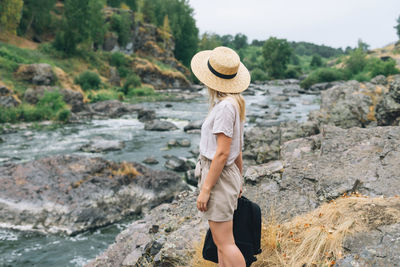 Young woman traveler in straw hat with backpack sits and looks at the mountain river, local travel 