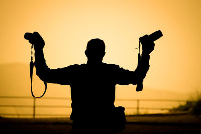 Rear view of silhouette man holding cameras against sky during sunset