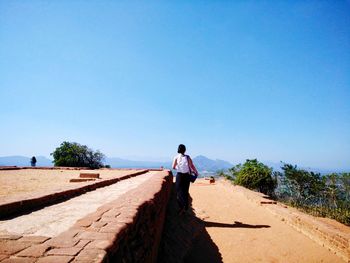 Rear view of man walking by retaining wall against clear blue sky