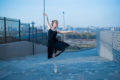 Portrait of young woman with arms outstretched against sky in city