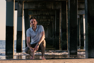 Full length portrait of man crouching below pier on shore at beach