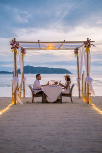 People sitting on chair at beach against sky during sunset