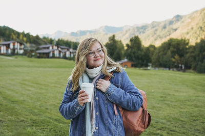 Young blonde woman in jeans coat with paper cup of morning coffee at the chalet cottage village 