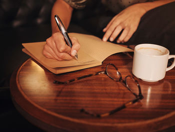 From above crop unrecognizable woman writing with pen in notebook at round wooden table with coffee and glasses in cafe