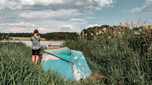 Rear view of woman standing by boat on grassy field against cloudy sky