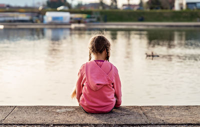 View from behind of sad abandoned little girl with braids sitting alone looking at lake outdoors