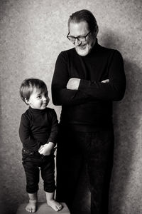 Grandfather with beard and glasses stands with his grandson in the studio near the wall