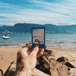 Cropped image of hand holding instant print transfer at sea shore against sky