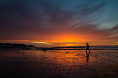 Silhouette man walking on shore at beach against orange sky