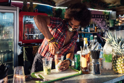 African american curly haired male bartender in mask preparing refreshing summer cocktail with lime and ice at counter of outdoor bar