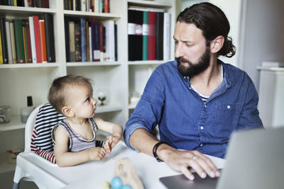 Mid adult businessman using laptop while looking at baby boy at home