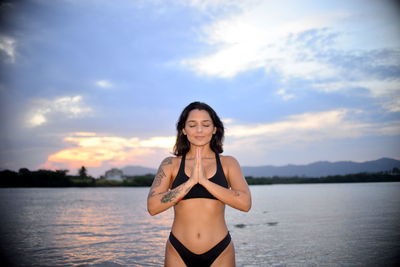 Young woman in bikini meditating by river against sky