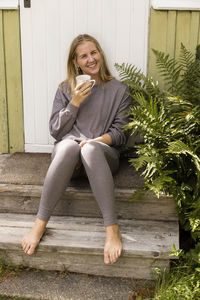 Portrait of happy woman holding mug while sitting on steps outside house