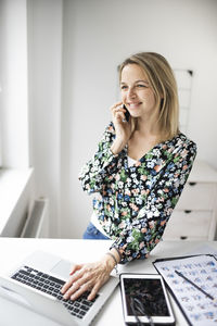 Smiling businesswoman talking on phone while using laptop in office