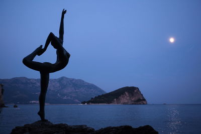 Silhouette of woman doing yoga on rock by sea against sky during dusk