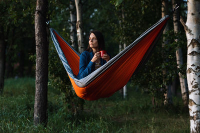 Female traveler is sitting in a hammock and drinking tea from a red cup. hiking outdoor recreation