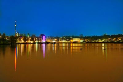 Illuminated buildings by lake against blue sky at night