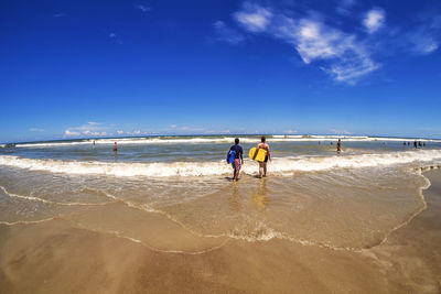 Rear view of people with surfboards walking at beach against blue sky on sunny day