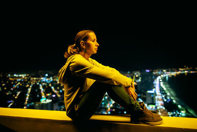 Side view of woman sitting on retaining wall against illuminated city at night