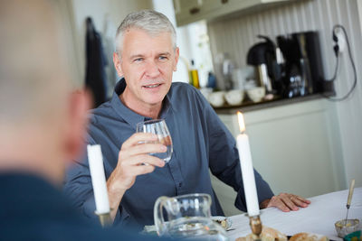 Mature man looking away while holding drinking glass on dining table with friend in foreground