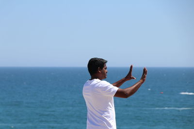 Rear view of mid adult man gesturing while standing at beach against sky