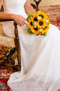 Low section of bride with bouquet sitting on chair at home during wedding ceremony