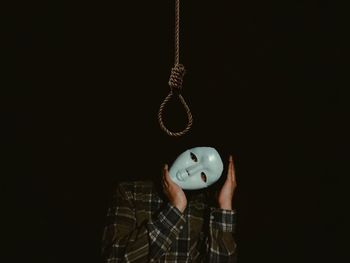 Portrait of person wearing mask standing below noose hanging against black background