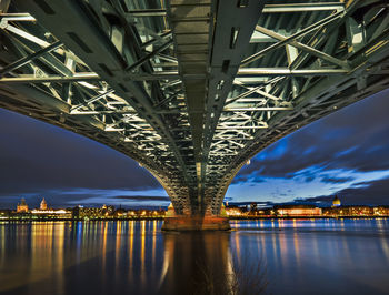Underneath view of illuminated bridge over river in city at dusk