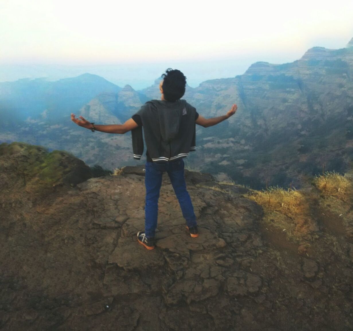 REAR VIEW OF MAN STANDING ON LANDSCAPE AGAINST MOUNTAINS