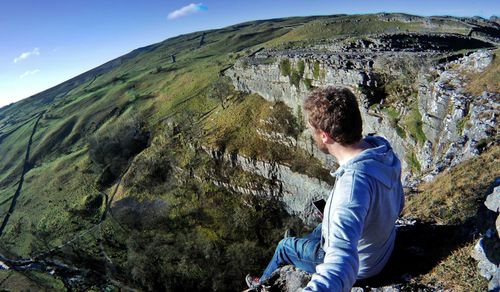 Side view of man sitting on cliff against sky