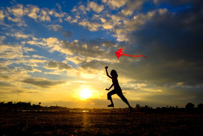 Silhouette girl flying kite while running on landscape during sunset