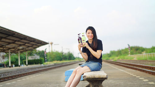 Portrait of woman holding bottles while sitting at railroad station platform