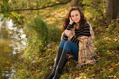 Full length of young woman smiling while sitting at grassy lakeshore