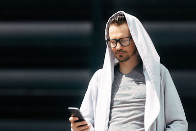 Man in hooded jacket texting on smart phone against black wall. copy space.