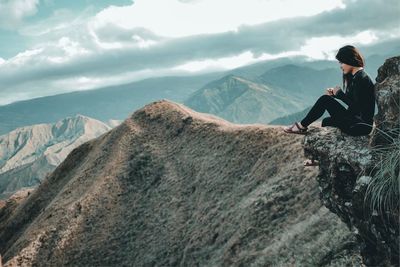 Side view of woman sitting on cliff against mountains