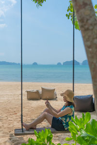 Young woman sitting on swing near beach and using smartphone to take photo during summer vacation