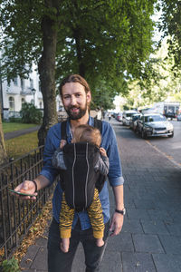 Portrait of happy man using mobile phone while carrying baby in carrier on sidewalk