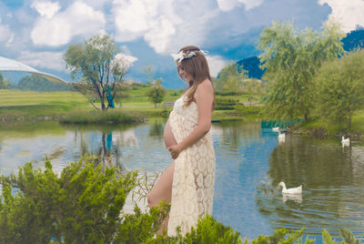 Side view of young woman standing by lake against sky