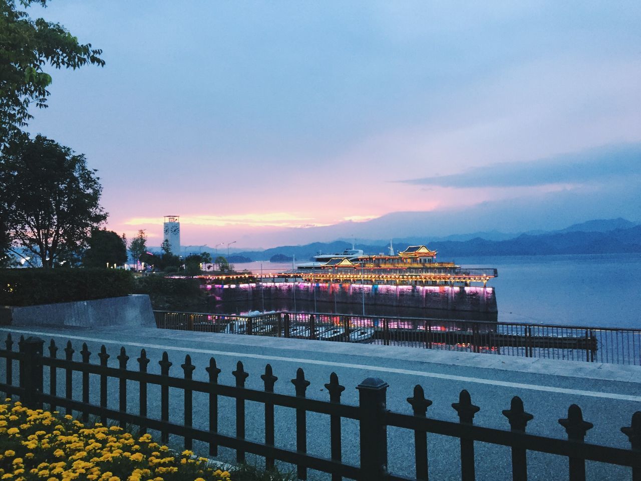 water, railing, tree, tranquil scene, sky, architecture, illuminated, tranquility, cloud, calm, bridge - man made structure, sea, scenics, blue, orange color, waterfront, nature, beauty in nature, outdoors, sun, majestic, no people, ocean