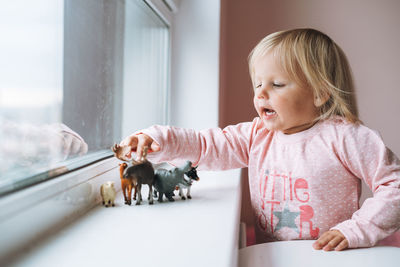 Little girl toddler playing with animal toys on window sill in children's room at home