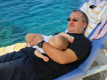 Man holding baby girl while lying on lounge chair against sea
